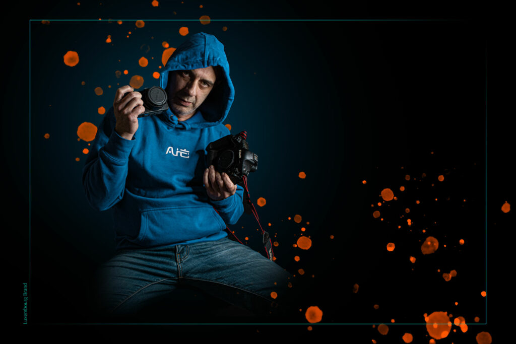 Imagen de un fotógrafo que sostiene 2 cámara en sus manos y está vestido con un hoodie azul y un pantalon azul y unas chispas como depintura al fondo en color naranja